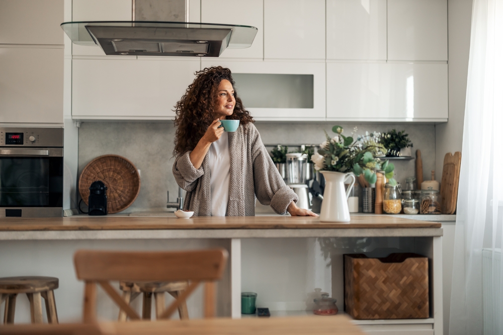A woman in a cozy robe stands in a bright kitchen with a morning coffee, relaxed and rested