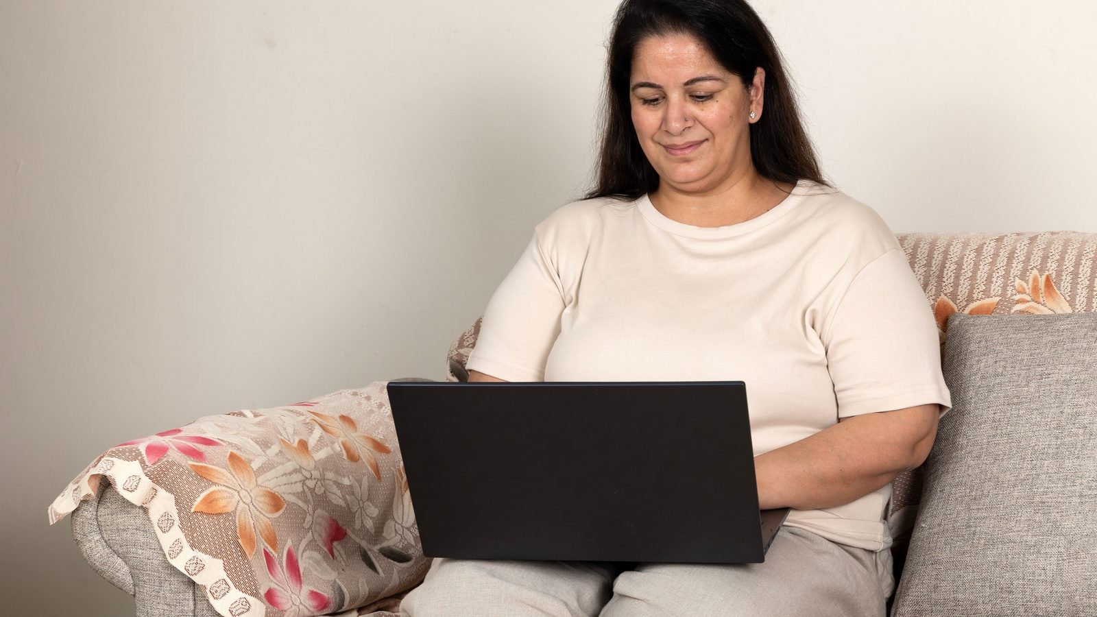 South Asian woman in her 40s sitting on a sofa at home using a laptop to compare HRT costs between telehealth and in-person care in Arizona