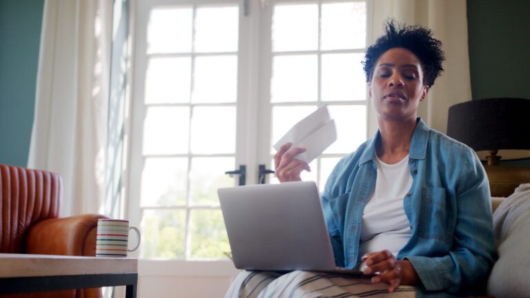 Black woman in her 40s fanning herself during a hot flash while working on her laptop at home, considering HRT or non-hormonal options like Veozah