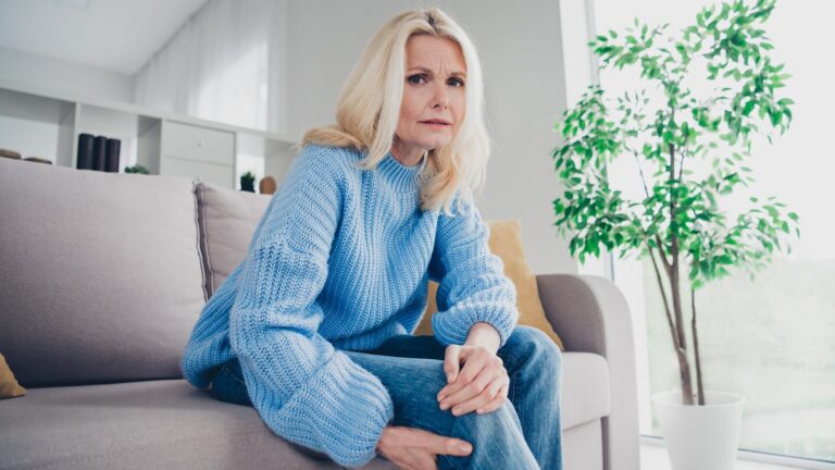 Woman in her 50s sitting on a sofa at home holding her knee, experiencing perimenopause joint pain